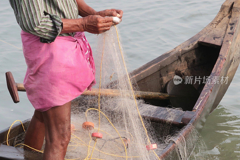 印度南部喀拉拉邦Kollam的渔民在小木渔船上，斜靠在白色尼龙渔网上，为海鲜市场捕捉小鱼。渔民穿着传统的粉红色纱笼/杜蒂捕鱼